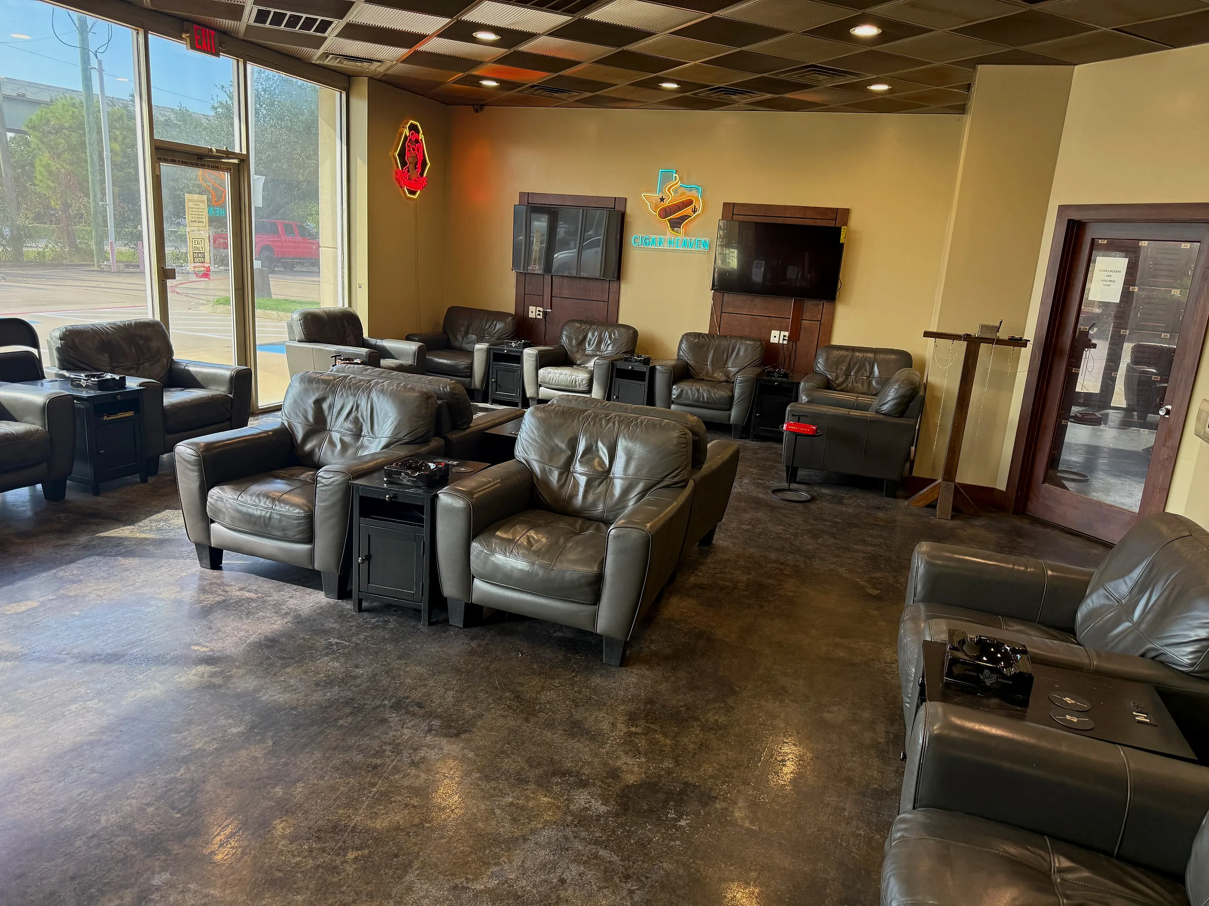 The lounge at Cigar Heaven — gray leather armchairs facing two TVs and the neon Cigar Heaven Texas sign, with the locker room door on the right.