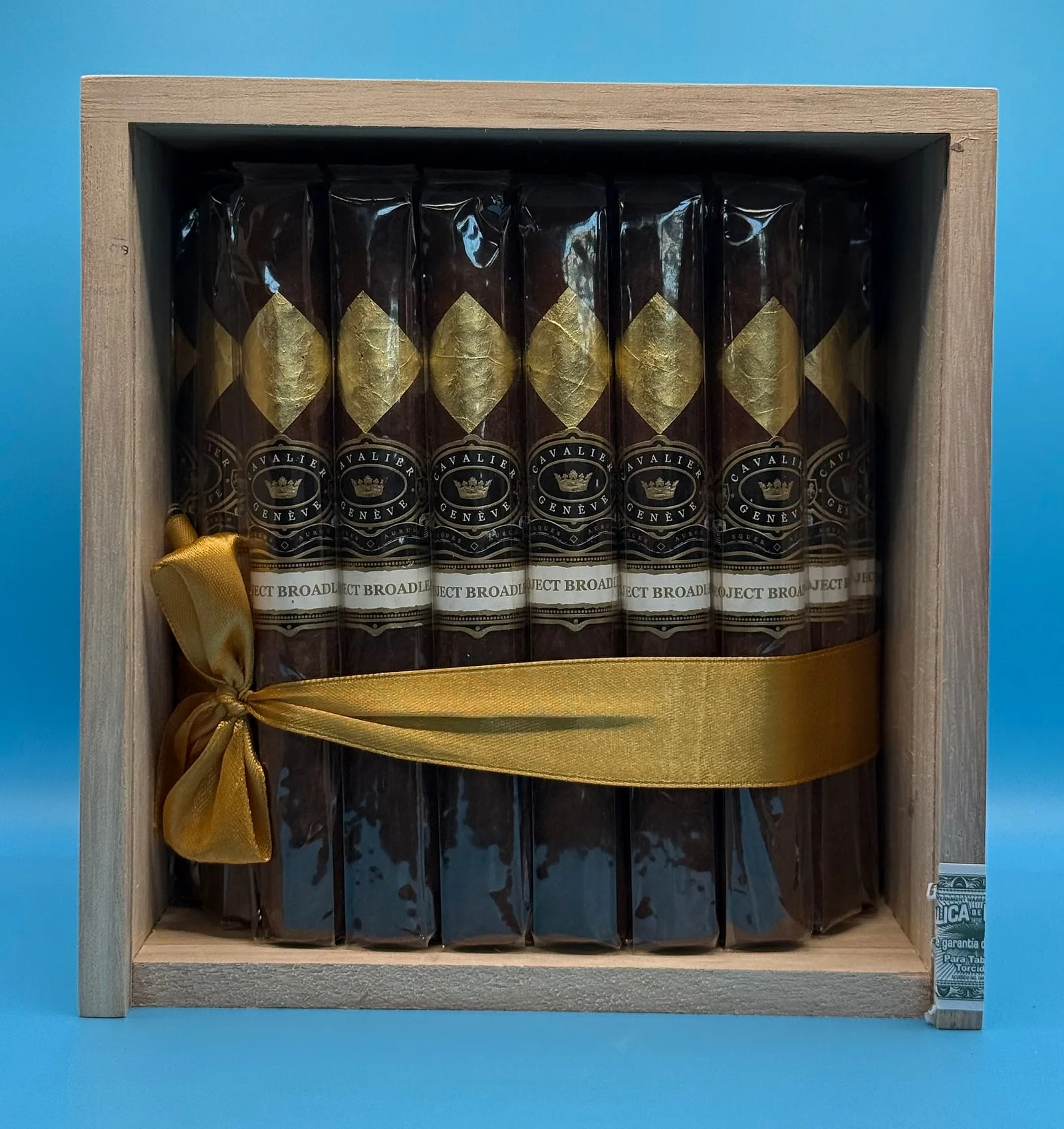 An open box of Cavalier Genève Project Broadleaf cigars with gold-leaf diamonds on the wrappers, tied with a gold ribbon.