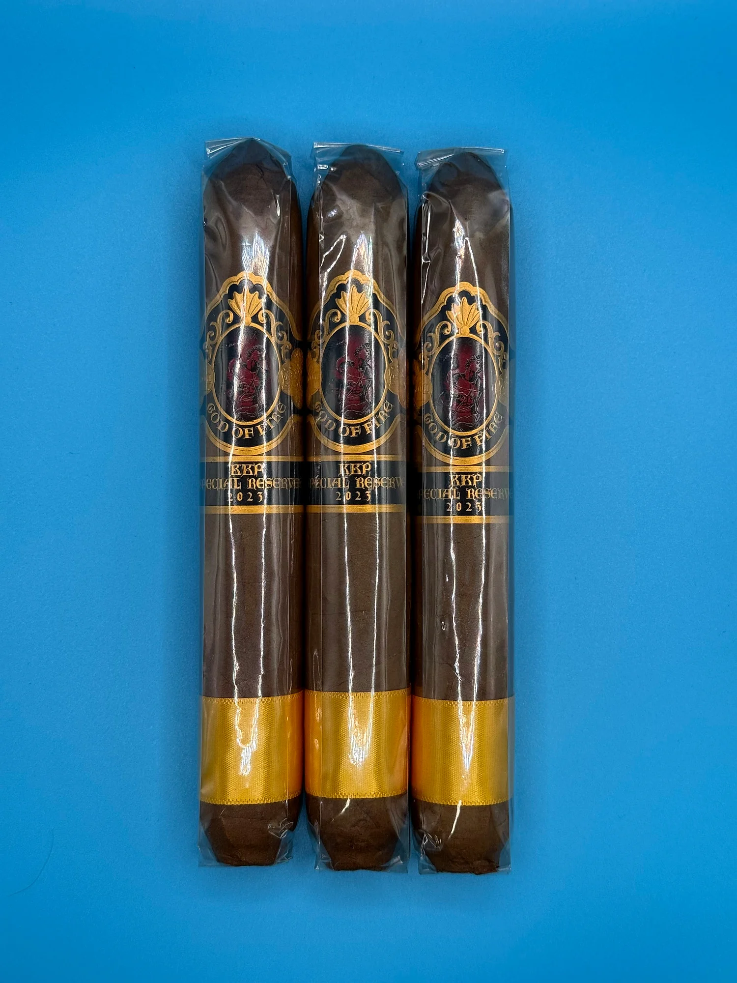 Three God of Fire KKP Special Reserve 2023 cigars in cellophane, with black-and-gold bands and gold ribbon at the foot, on a blue background.