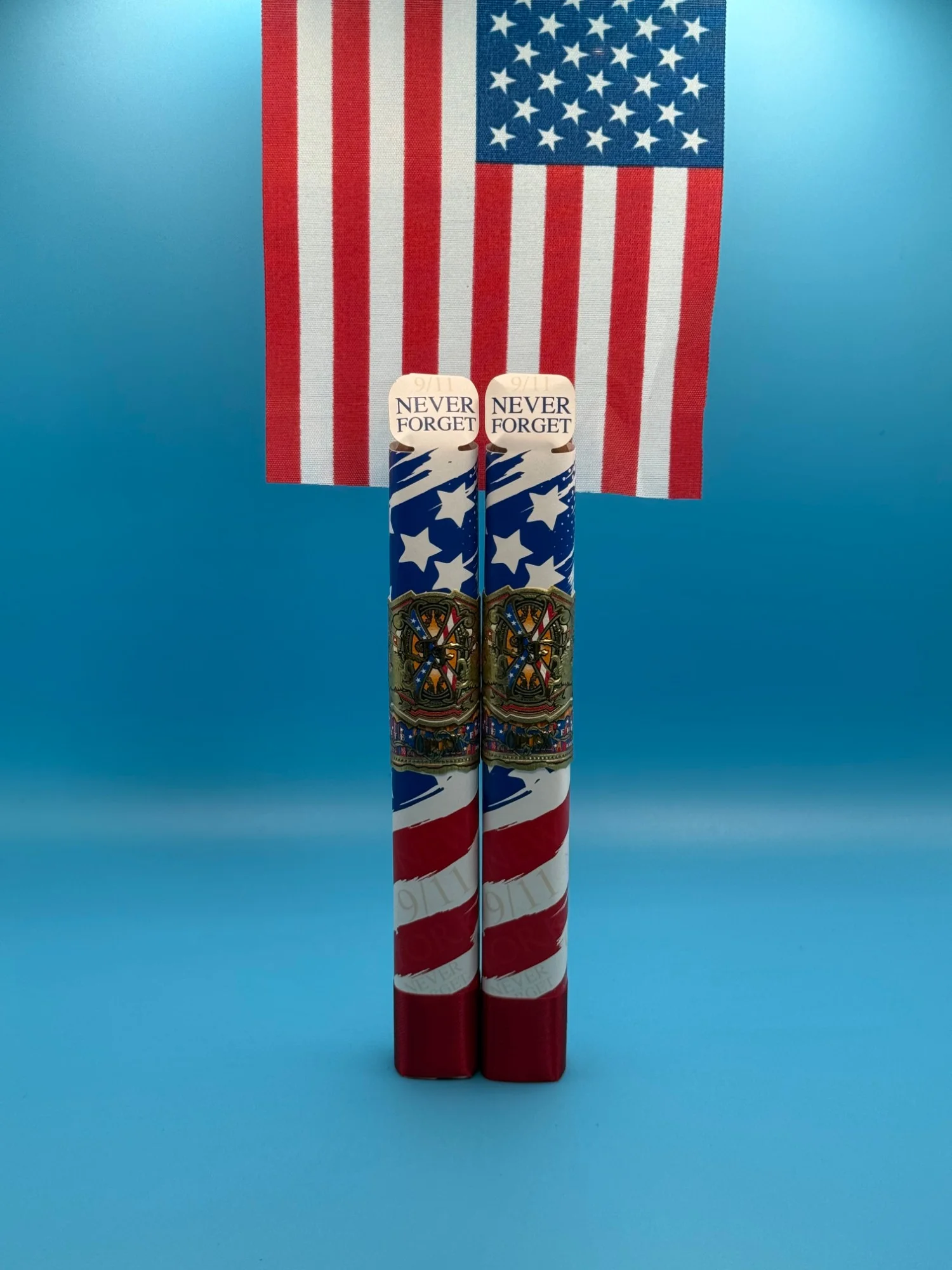 Two Fuente Fuente OpusX Rexilient cigars in 9/11 “Never Forget” commemorative stars-and-stripes wraps, standing in front of an American flag.