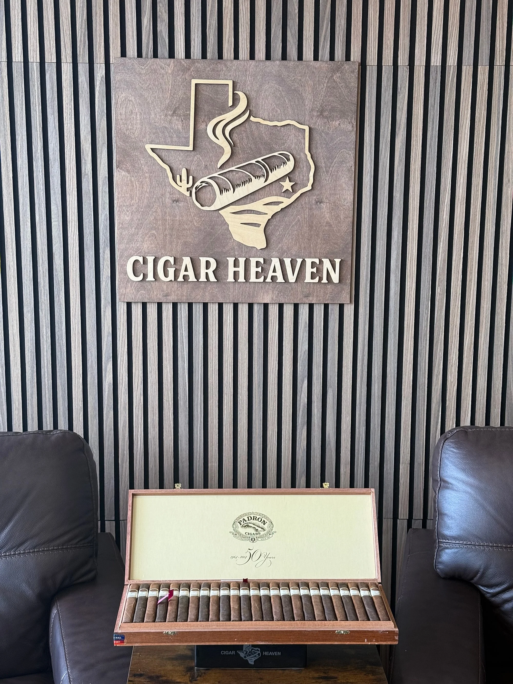 An open box of Padrón 50th Anniversary cigars, marked 1964–2014 50 Years, with Maduro and Natural wrappers side by side, below the Cigar Heaven sign in the lounge.