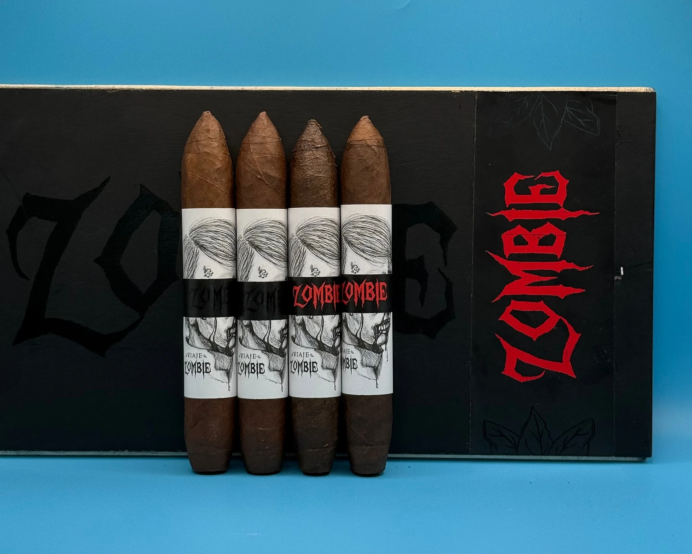 Four Viaje Zombie cigars with sketched zombie artwork bands, two in black and two in red, against a black Zombie box.